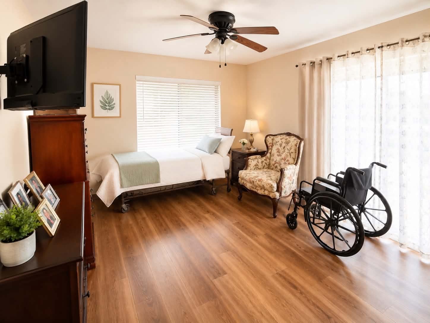 A spacious private bedroom at Tessa Place 2 with an adjustable bed, an upholstered armchair, family photos, and a wheelchair ready beside the bed.